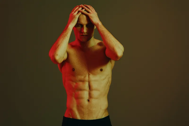 Tensed man with bare torso expressing frustration in a dramatic red light setting, showcasing raw emotions and intensity in a striking composition.