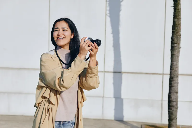 Photography, camera, outdoor, woman, smiling, casual, lifestyle, daylight, a candid moment of a smiling woman holding a camera outdoors beside a white wall, capturing spontaneous urban vibe and