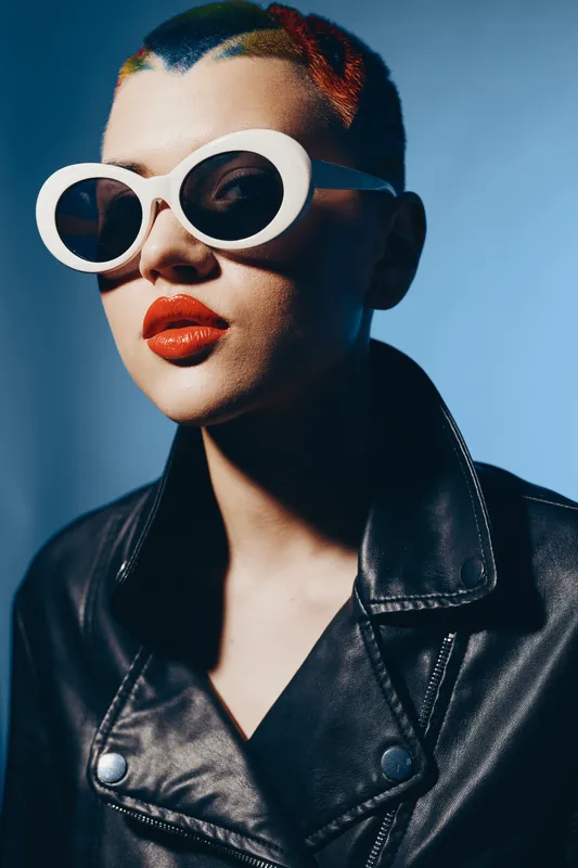 woman with bold red lipstick wearing white sunglasses and black leather jacket, modern edgy style with dramatic lighting and blue background