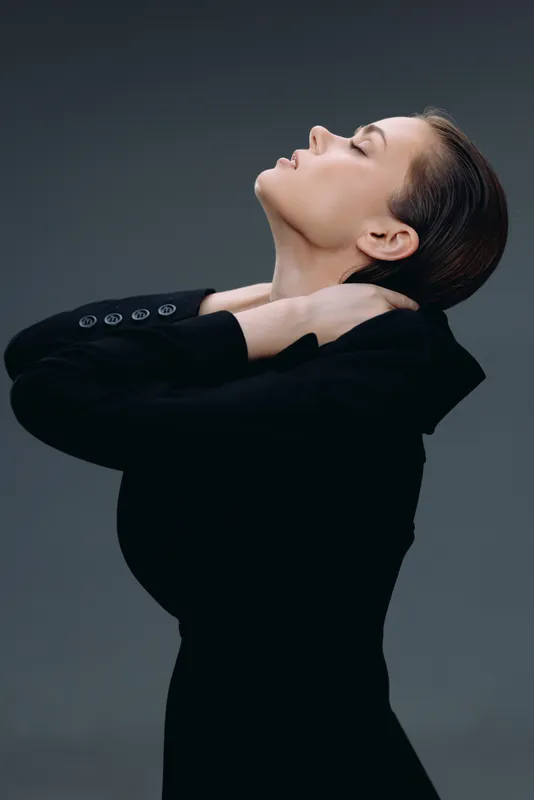 Profile of a woman in black outfit touching her neck with closed eyes, showing elegance and calmness with a dark gray background.