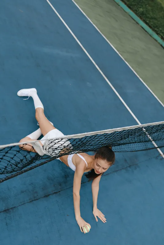 Young female tennis player stretching to catch a ball on the court, showcasing determination and athleticism in sporty attire with a vibrant blue background