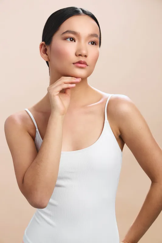 Asian woman in a white tank top stands against a soft beige backdrop, posing thoughtfully with calm confidence and poised expression, ideal for lifestyle and fashion contexts.
