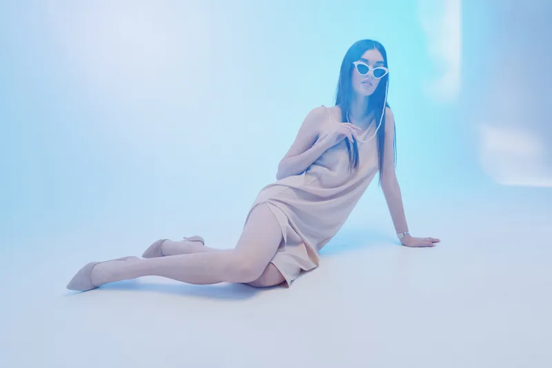 Fashionable woman in a beige dress and sunglasses poses on the floor in a minimalistic studio with soft blue lighting. Concept for beauty, fashion, and modern style.