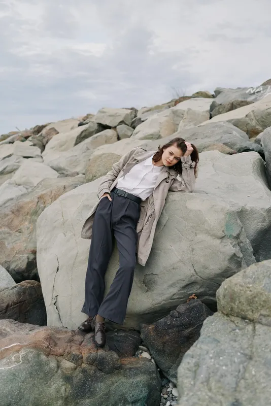 Young woman in casual outfit leans on large rocks outdoors under cloudy sky. Concept of fashion, nature, relaxation, and autumn style in natural environment.