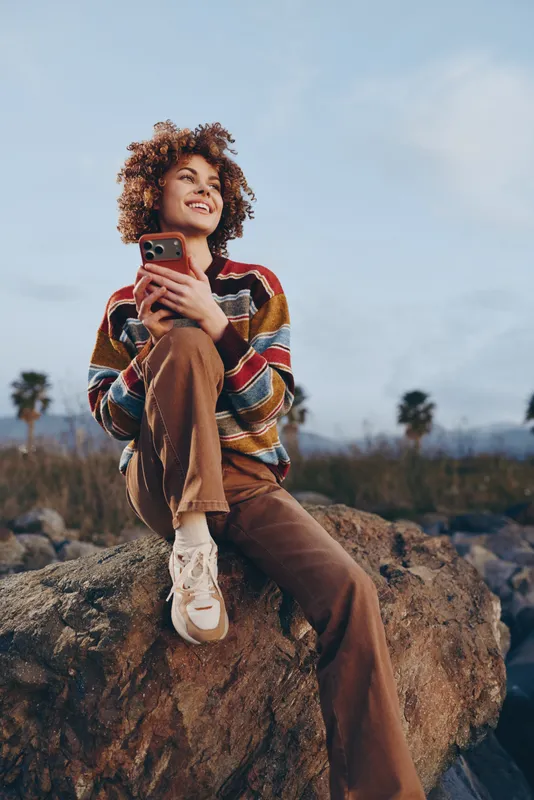 Woman lifestyle outdoors, a young woman sitting on a rock, curly hair framing her face, holding a smartphone, casual striped sweater and jeans, soft sunlight, landscape.
