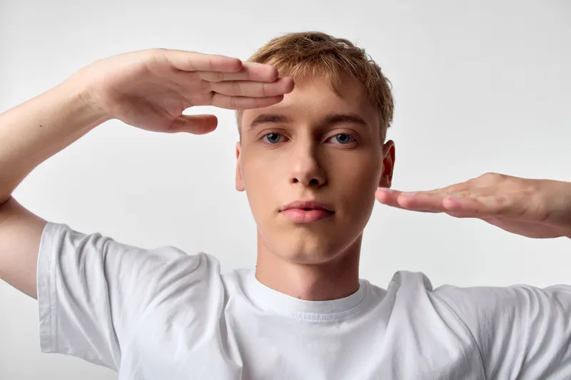 Framing a shot with hands around the face, a young man in a white t shirt demonstrates photography composition, balance and focus in a clean studio setting