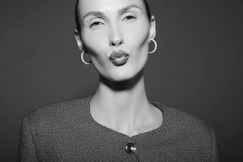 Portrait of a light skinned Caucasian woman with earrings blowing a kiss, wearing a textured jacket against a dark background. Concept of beauty, fashion, and affectionate gesture.