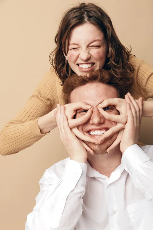 Happy young couple having fun together, woman playfully covering man's eyes with her hands. Both smiling and enjoying a light moment in casual clothing on beige background.