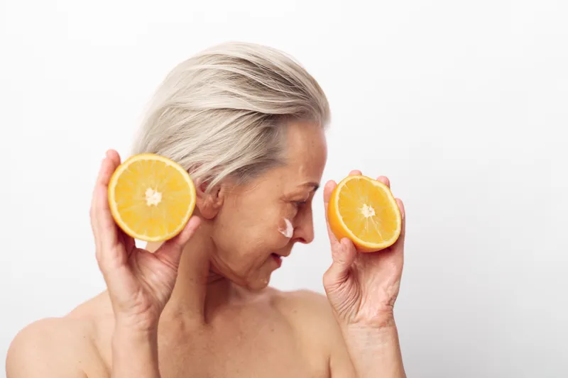 Mature woman with silver hair holding two orange halves near her face on white background. Concept of natural beauty, skincare, wellness, and healthy lifestyle for aging skin care.