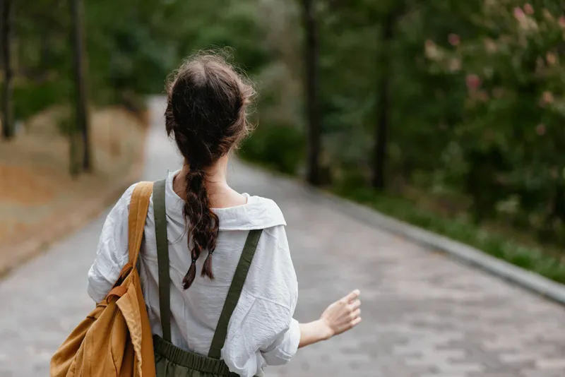 A young woman walks through a serene park path in casual attire, expressing a relaxed, carefree emotion with a natural backdrop in soft greens and browns