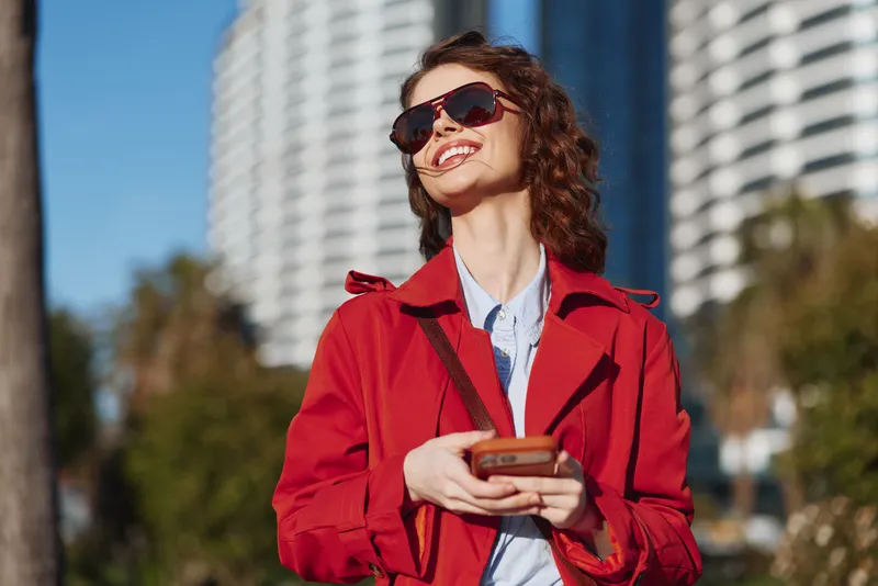 A young woman in a red coat smiles while checking her phone outdoors in an urban park on a sunny day, with oversized sunglasses reflecting the bright cityscape, capturing a modern lifestyle moment.