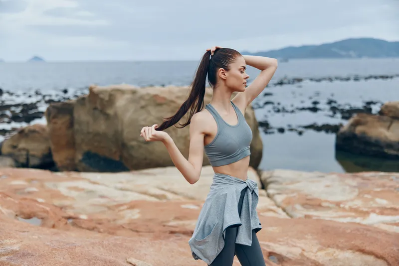 Fit young woman in sportswear enjoying nature by the sea, showcasing a healthy lifestyle and physical fitness, with a tranquil coastal background and natural colors