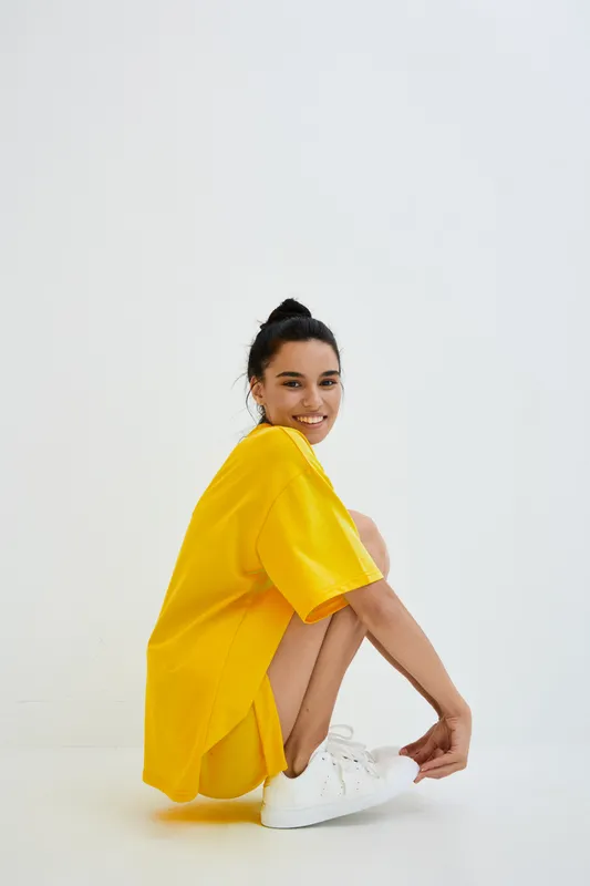 Smiling woman wearing a bright yellow oversized t-shirt and white sneakers, sitting in a relaxed pose against a plain white background with natural lighting.