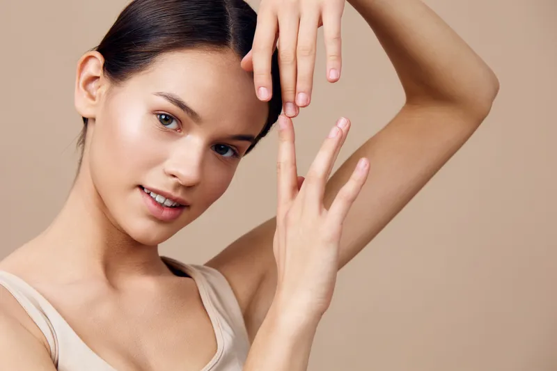 Young woman with smooth skin posing gracefully with hands, wearing a light tank top, embodying beauty and tranquility against a warm beige background