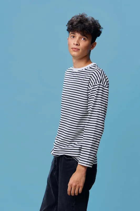 young man in striped shirt standing against blue background with casual posture and neutral expression, showcasing modern street style and youthful vibe