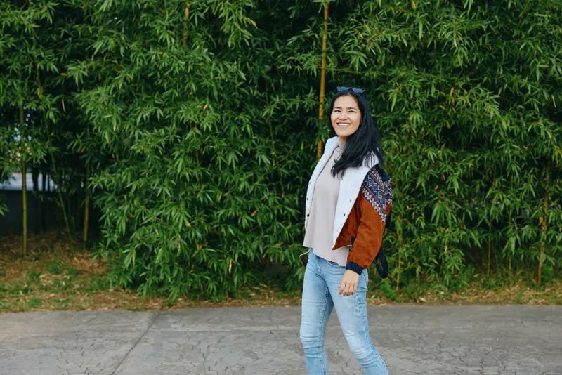 Woman walking outdoors along a green hedge, smiling, wearing a patterned cardigan and scarf, casual denim jeans and a bag, a cheerful natural street style moment