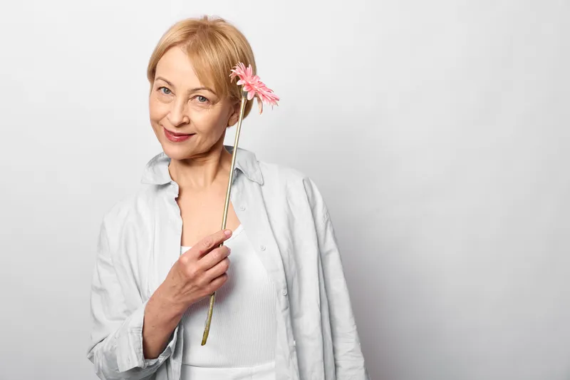Older woman in a white medical coat holds a pink flower, smiling in studio portrait, confident healthcare mood, soft light, isolated on clean white.