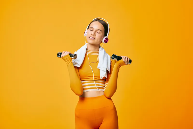 Fitness scene with a woman in bright orange activewear smiling and holding dumbbells, a towel around her neck and headphones on, portraying a lively workout mood against a vibrant orange background.