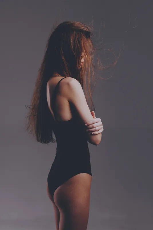 Profile of a thoughtful woman in black bodysuit with long hair and gray background, moody atmosphere.