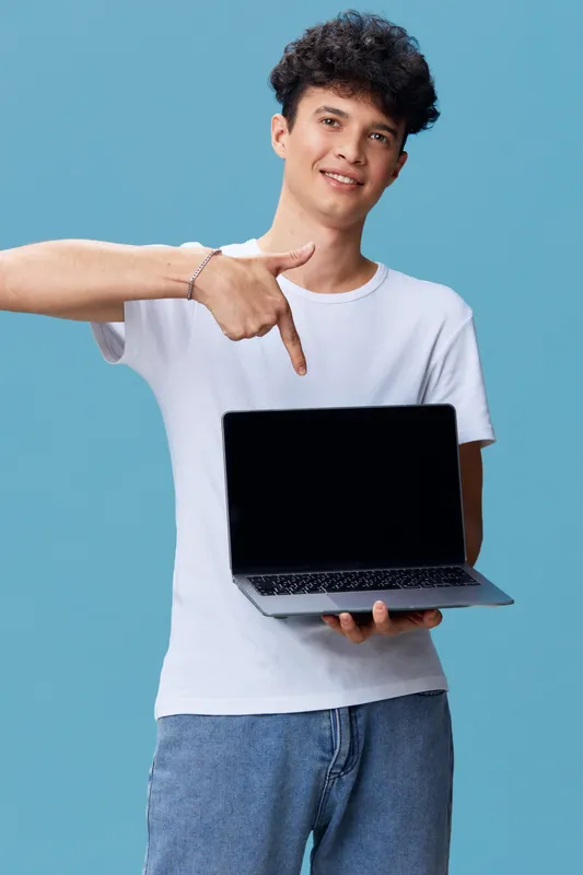 young man holding laptop pointing at screen, casual clothing, curly hair, blue background, technology, smiling, digital device