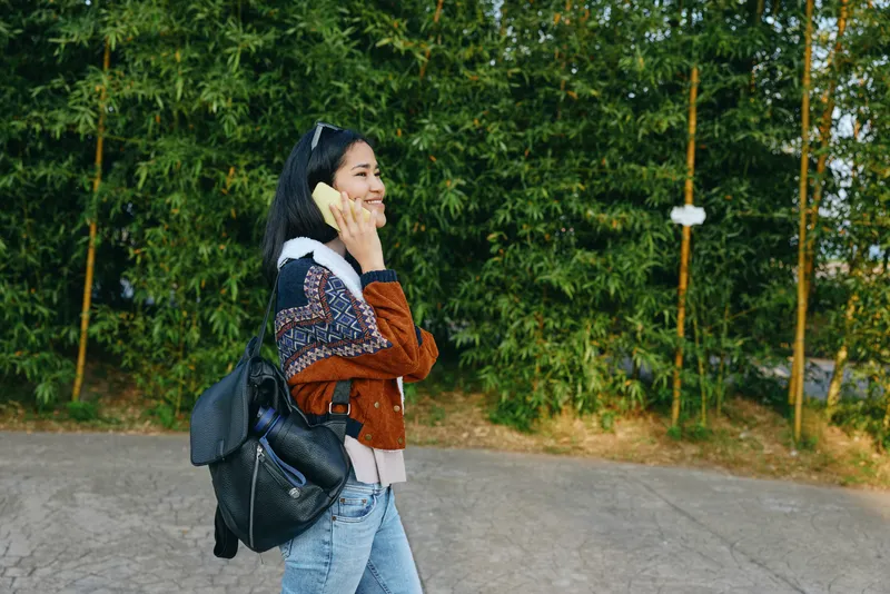 Young woman walking outdoors, talking on a phone while carrying a backpack, casual denim jacket and jeans, street scene with green trees, candid lifestyle moment during errands