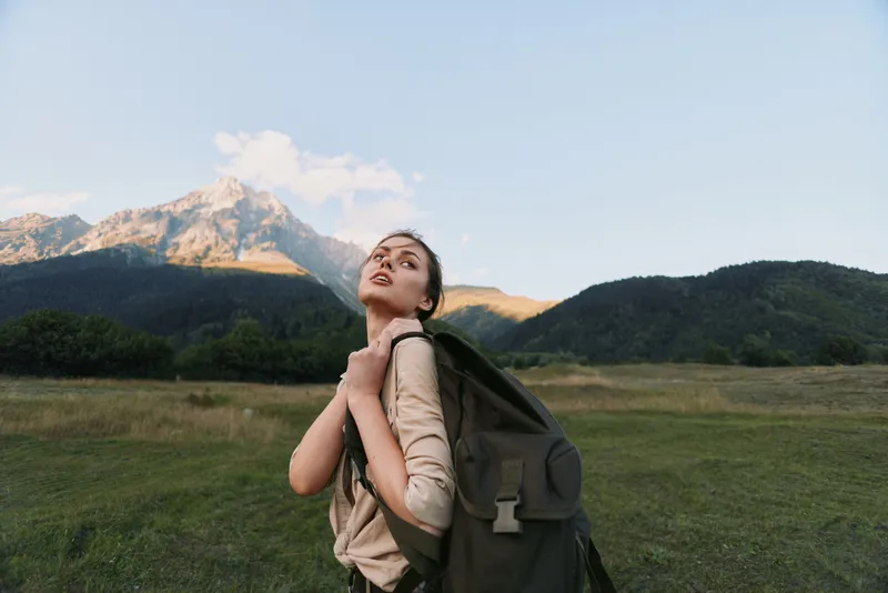 Young woman with a backpack gazes upward in an open meadow, mountains in the distance. Outdoor lifestyle, exploration, and self discovery through hiking journey.
