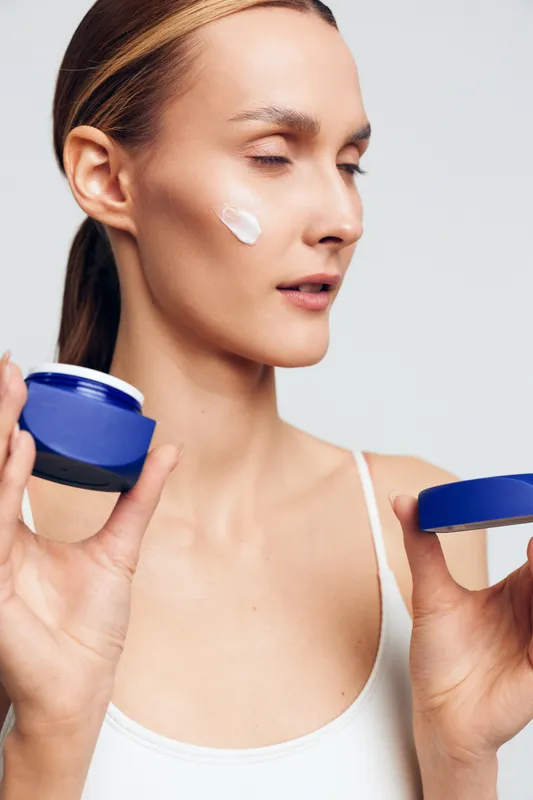 Woman applying face cream on cheek with eyes closed, holding open jar in hand. Skincare routine, healthy skin, moisturizing concept with soft natural lighting on white background.