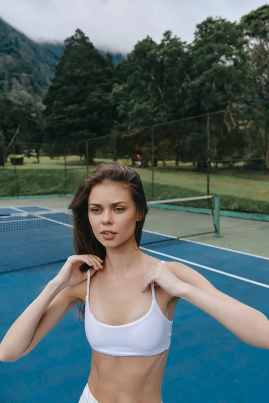 Young woman in a white sports bra showcasing athleticism on a tennis court surrounded by greenery, reflecting fitness and a healthy lifestyle