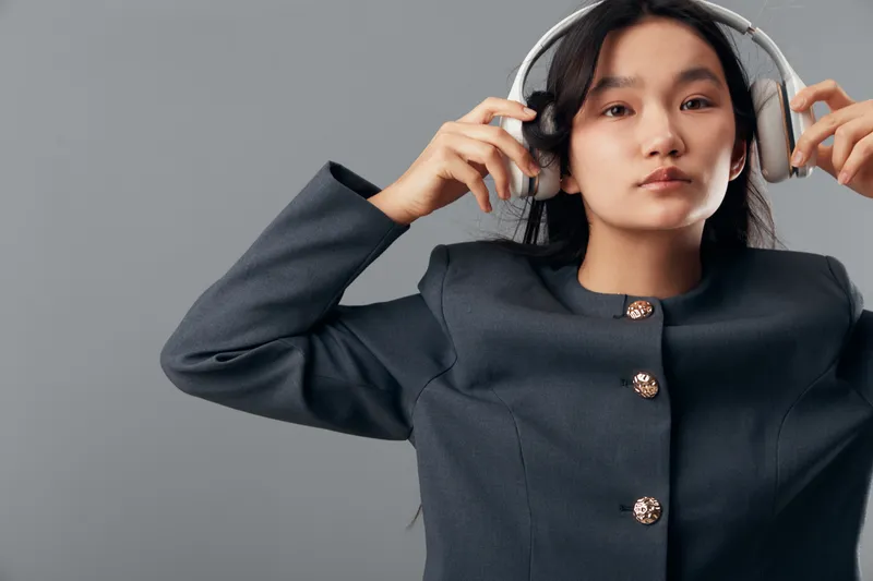Young woman wearing headphones and fashionable jacket, posing against gray background with neutral expression in studio for music, technology, and style concept.