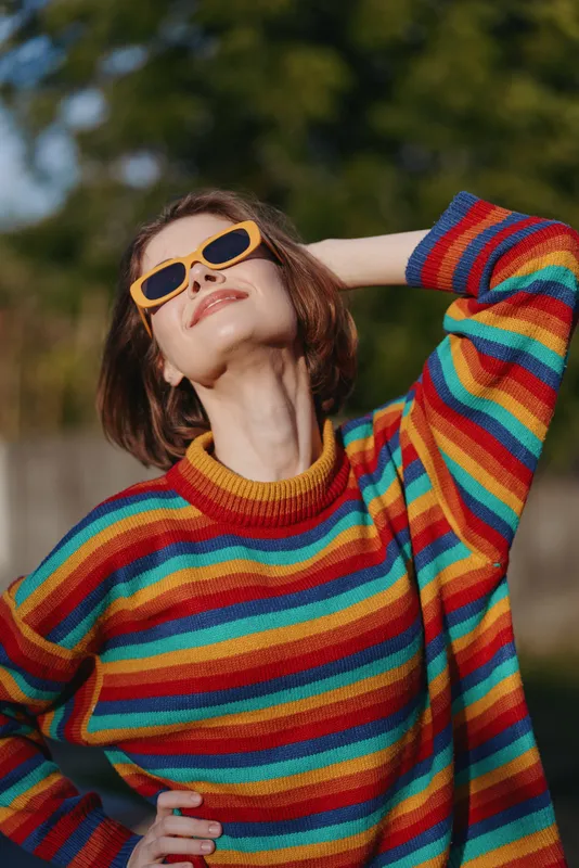 Woman, rainbow sweater, sunglasses, smiling, outdoors, casual fashion — Young woman in her 20s with bob haircut wearing yellow sunglasses and a striped sweater, posing carefree in sunlit park.