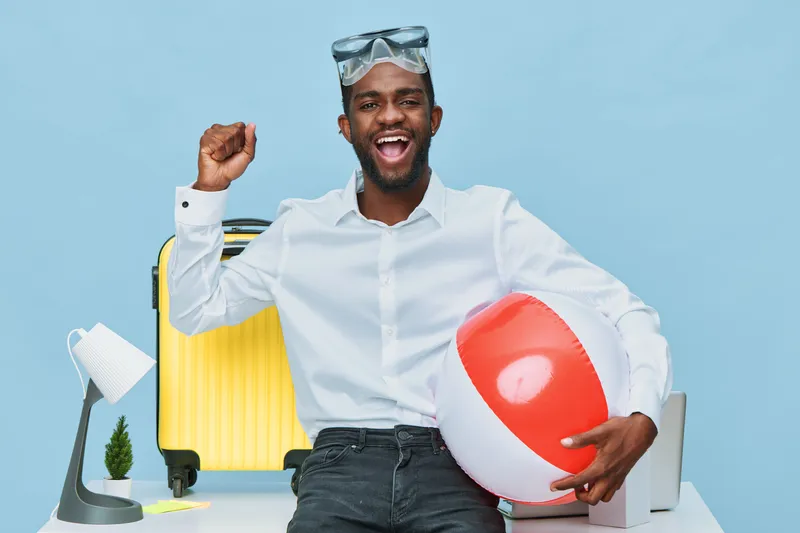 Smiling man with dark skin celebrating summer vacation, wearing glasses and casual white shirt, holding a large beach ball, surrounded by travel and leisure items in bright studio setting. People life