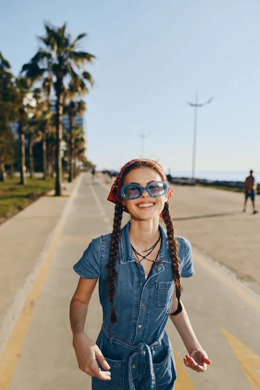 Lifestyle woman in modern boho-western denim street style smiling confidently outdoors with sunglasses and braided hair, vibrant film color capturing sunny day energy.