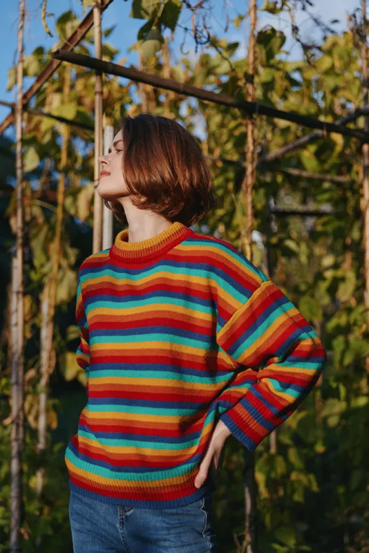 Woman in colorful striped sweater portrait outdoors, short hair and casual pose in garden sunlight, side profile fashion shot conveying cozy knitwear, relaxed mood and autumn warmth.