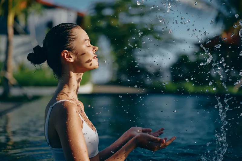 Young woman outdoors under a gentle shower by a pool, water droplets glisten on sunlit skin, celebrating refreshment, wellness, and a carefree lifestyle in a bright, serene moment.