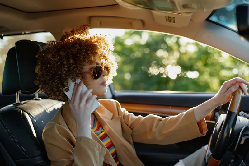 Young woman with curly hair wearing sunglasses and a beige coat drives a car, exuding retro film vibes and confident street style in bright afternoon light.