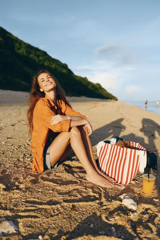 Young woman with long curly hair sits on a sandy beach wearing an orange shirt and denim shorts while enjoying a sunny day near a striped beach bag and a drink.