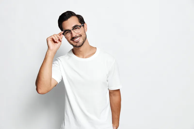 Smiling man wearing glasses poses confidently while adjusting his eyewear with a cheerful expression on his face against a solid white background. Lifestyle concept