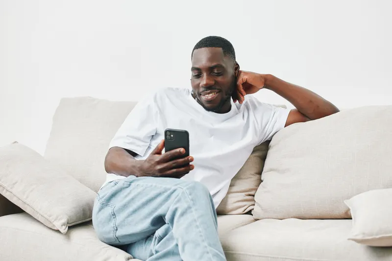Smiling young man relaxing on a couch while browsing on his smartphone, showcasing a casual lifestyle in a minimalist interior design