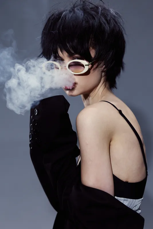Stylish woman wearing sunglasses exhaling smoke in a dark moody setting with black outfit and short hair.