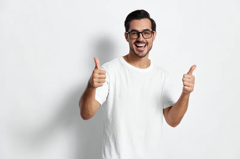 Cheerful smiling young man showing thumbs up wearing glasses and a white t-shirt against a plain light background. Positive lifestyle concept.