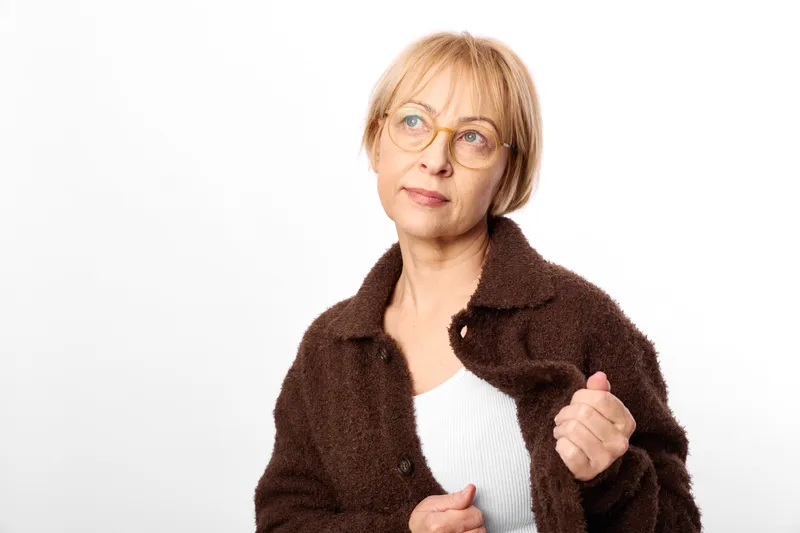 Mature woman with glasses, brown fur jacket, studio portrait on white background, slight smile, hands relaxed, calm confident expression, looking at camera
