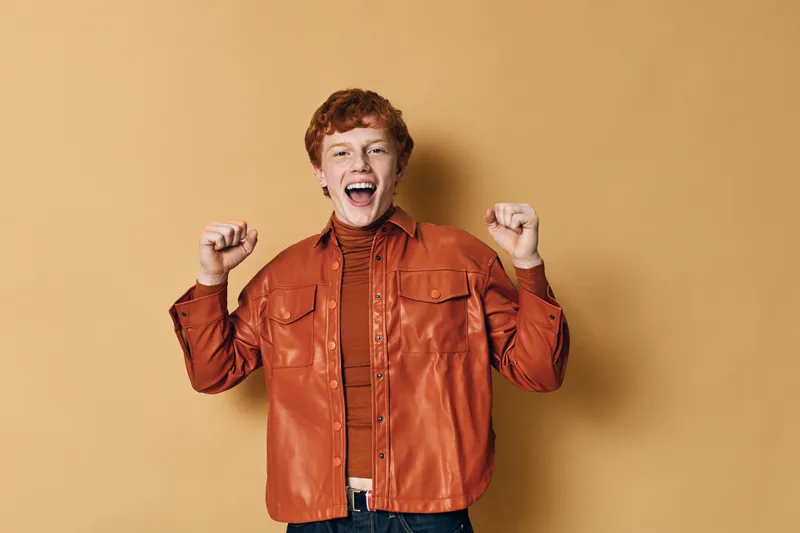 Young man with red hair wearing orange jacket and striped shirt smiling and raising hands excitedly for celebration on beige background indoors studio.