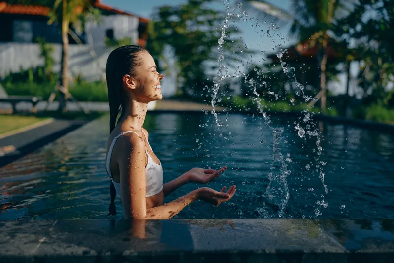 Young woman with long braided hair enjoys playful water splashes while sitting in a swimming pool on a sunny day, expressing relaxation and happiness in a casual white swimsuit.