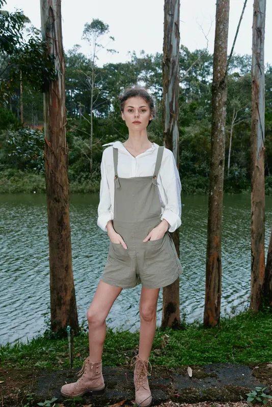 Young woman in overalls poses confidently by a serene lake, surrounded by lush trees, expressing a sense of adventure and connection to nature