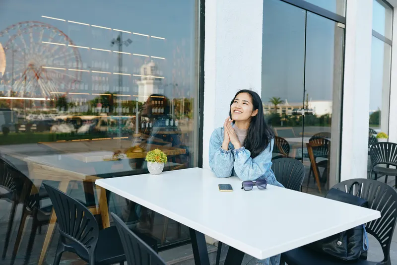 A thoughtful woman sits at an outdoor cafe table, sunlight on glass, reflecting urban life and a calm moment of quiet dining and relaxation