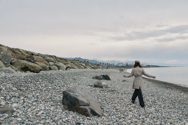 Woman in beige coat and hat walks along rocky beach under cloudy sky. Calm sea and distant mountains create peaceful atmosphere. Concept of solitude, nature, and autumn outdoors.