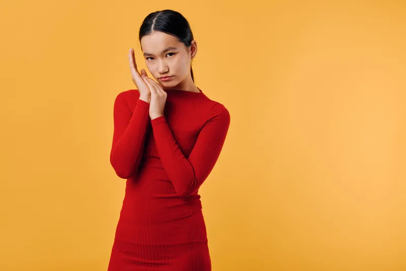 Young woman wearing red dress posing with hands near face against yellow background. Fashion portrait, stylish outfit and confident expression with clean composition and bright color contrast.