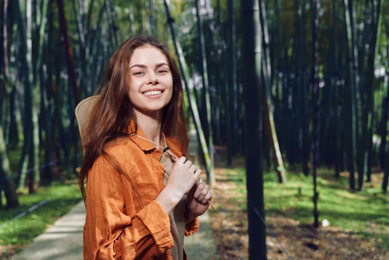 Woman smiling on forest path with backpack and portrait pose, nature outdoors scene with sunlight through trees, casual jacket, hiking travel mood and happy youthful energy