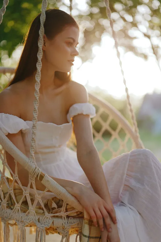 Serene young woman in white dress sitting on a swing, basking in golden sunset light with dreamy emotions, nature background