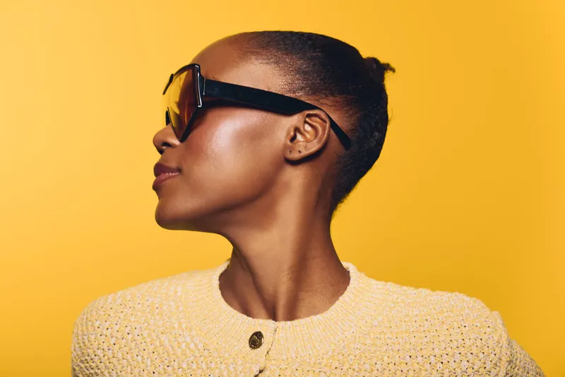 Profile of a confident black woman wearing large sunglasses and a yellow sweater against a yellow background. Concept of fashion, style, African ethnicity, and modern portrait.